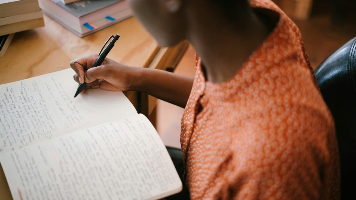 Focused black woman writing in a notebook with stacked books on a desk, illustrating learning.