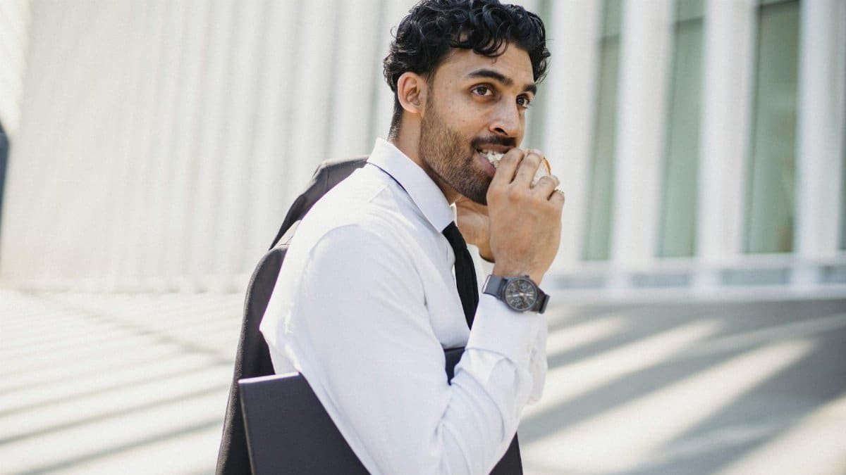 Smiling businessman in corporate attire enjoying a quick snack outdoors in a modern urban setting.
