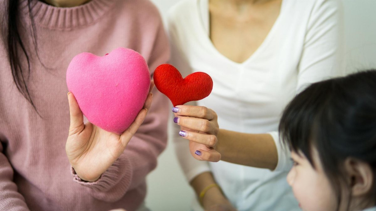 Unrecognizable ladies in casual wear demonstrating decorative hearts while sitting on couch near small kid in light room in daylight