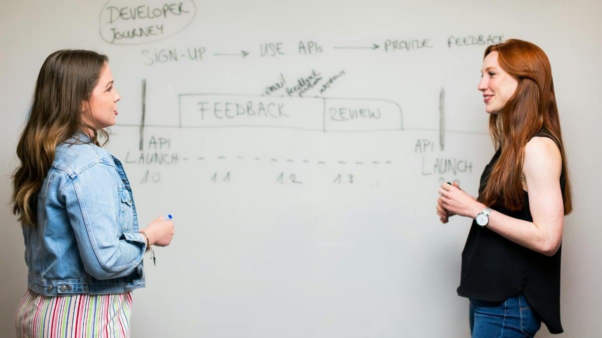 Two women engaging in a discussion about API development processes at a whiteboard.