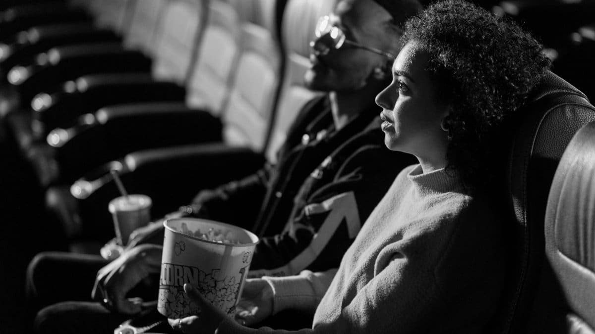 A couple sits together watching a movie in a classic black-and-white cinema setting.