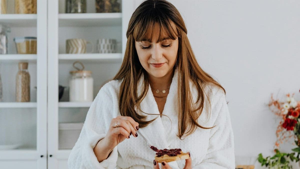 Woman in a bathrobe spreading jam on toast in a bright kitchen.
