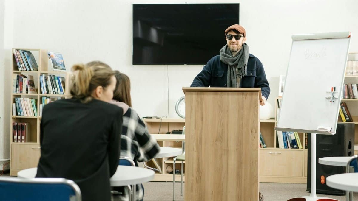 A man with sunglasses conducts a presentation on Braille in a library setting, engaging attentive listeners.