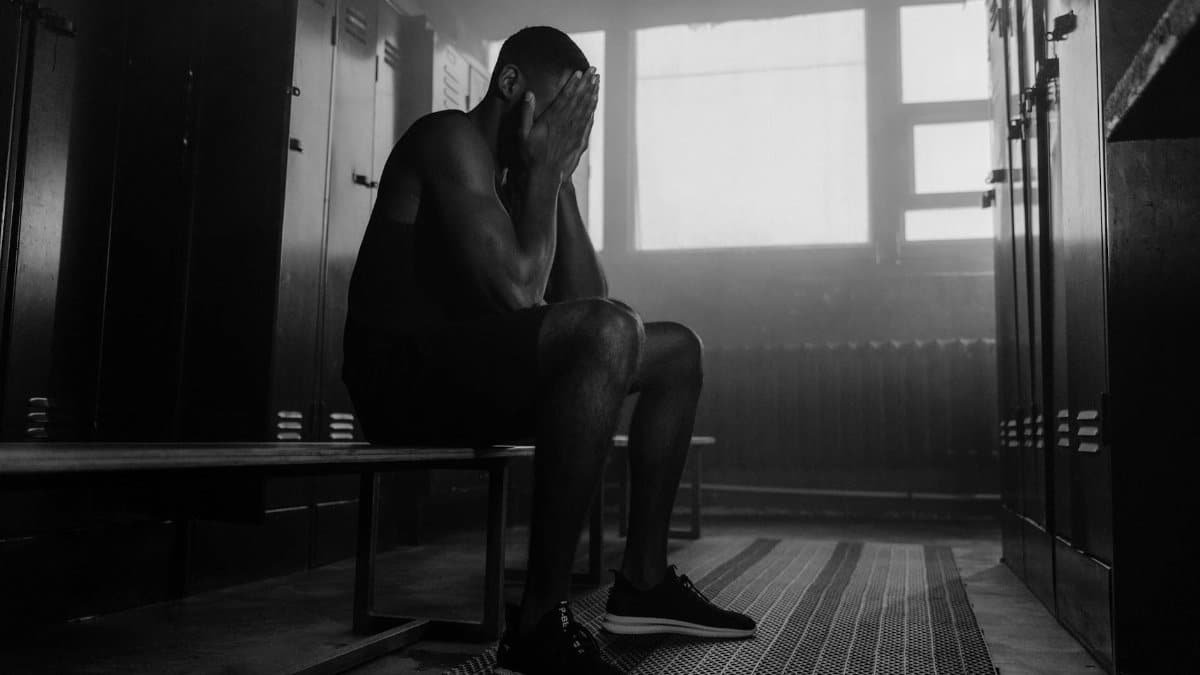 Monochrome image of a man sitting in a locker room with his face in hands, conveying contemplation or stress.