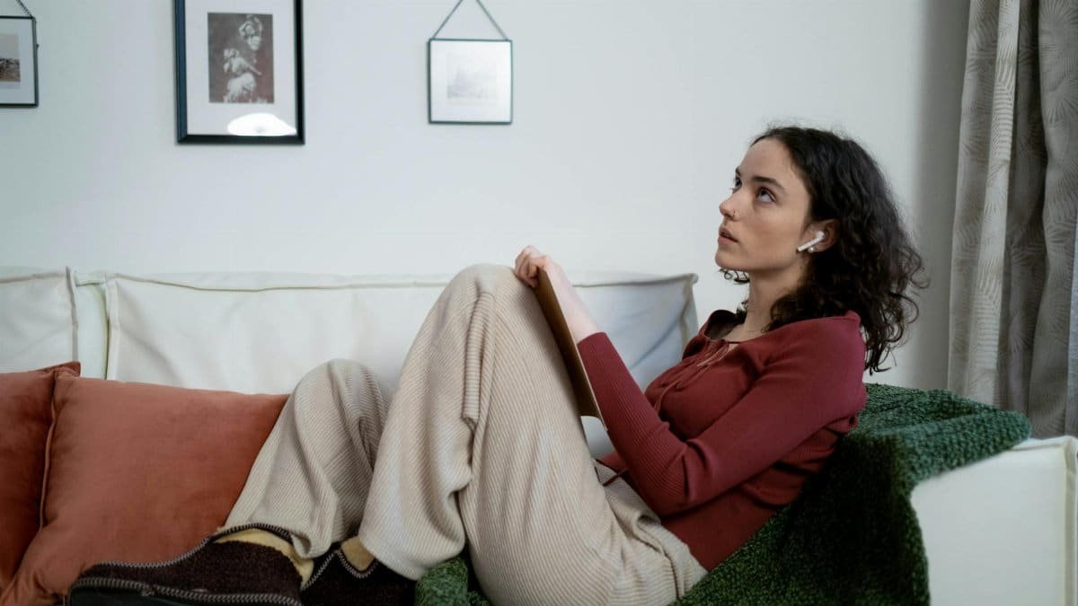 A young woman with curly hair sits on a sofa wearing AirPods, appearing deep in thought.