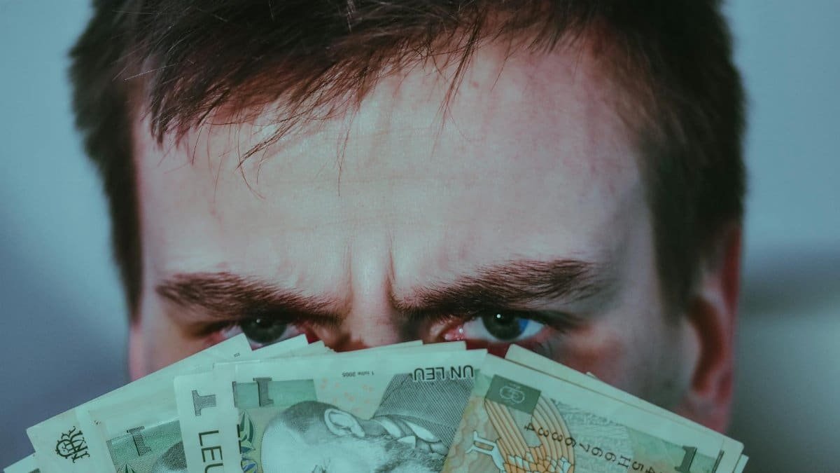 A man hides his face behind banknotes, conveying financial stress indoors.