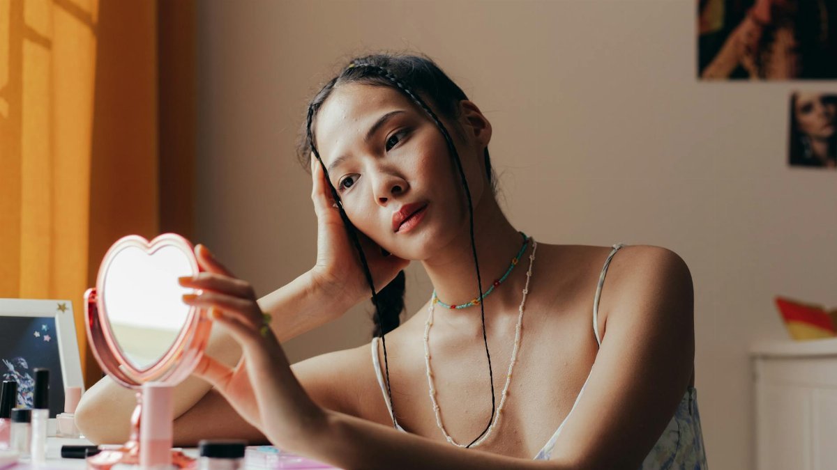 Elegant woman with braided hair gazing into a heart-shaped mirror in a warmly lit room.