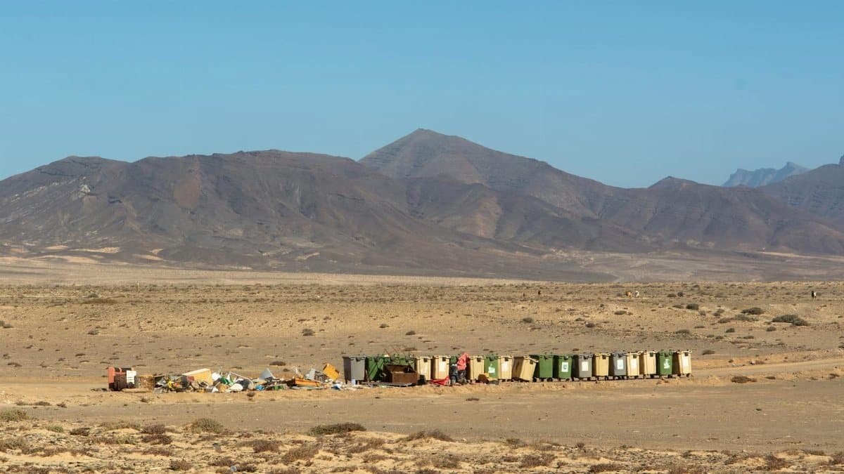 A row of recycling bins and waste in a desert landscape with mountains in the background.