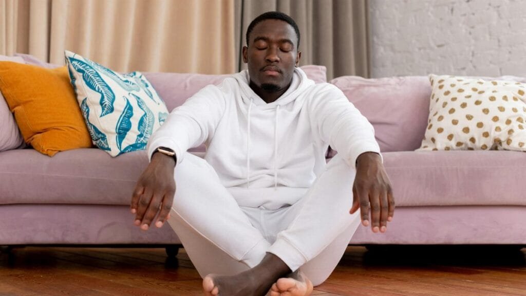 Calm African American male in white clothes sitting with closed eyes and crossed legs on floor against comfortable sofa in living room