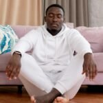 Calm African American male in white clothes sitting with closed eyes and crossed legs on floor against comfortable sofa in living room