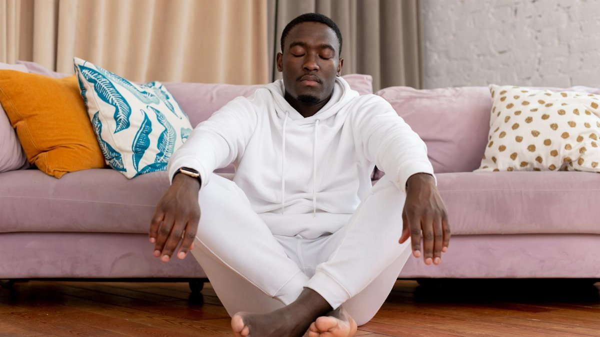 Calm African American male in white clothes sitting with closed eyes and crossed legs on floor against comfortable sofa in living room