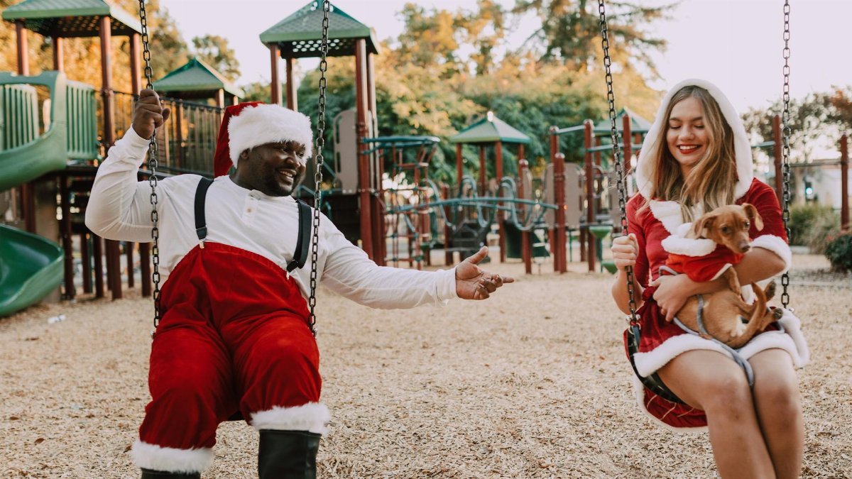 Two friends in Santa costumes enjoying a playful swing with a dog in a festive outdoor playground.