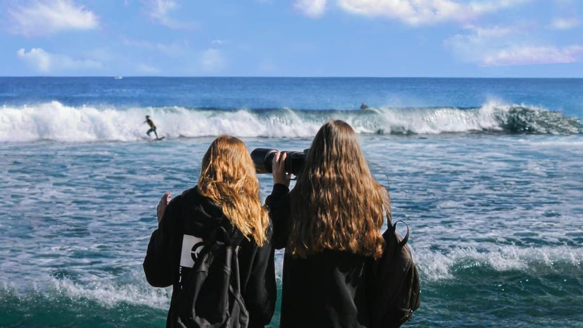 Two women photographing a surfer riding waves at the ocean, capturing the vibrant seascape.