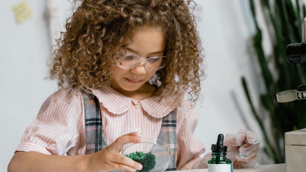 A young girl in glasses conducting a science experiment with green liquid indoors.