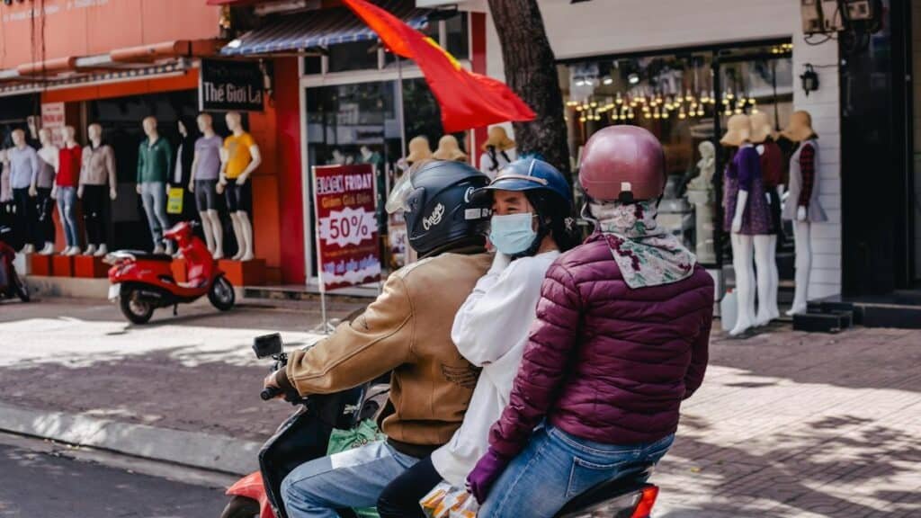Three people on a motorcycle ride through a bustling city street lined with shops and mannequins.