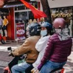 Three people on a motorcycle ride through a bustling city street lined with shops and mannequins.
