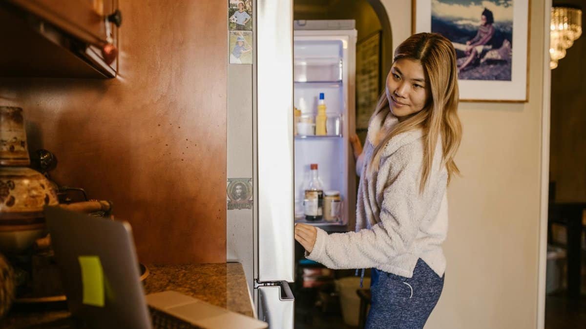 A woman at home checks her refrigerator while working on a laptop in a cozy setting.