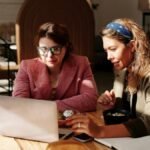 Two women working together on a laptop in a cozy café setting.