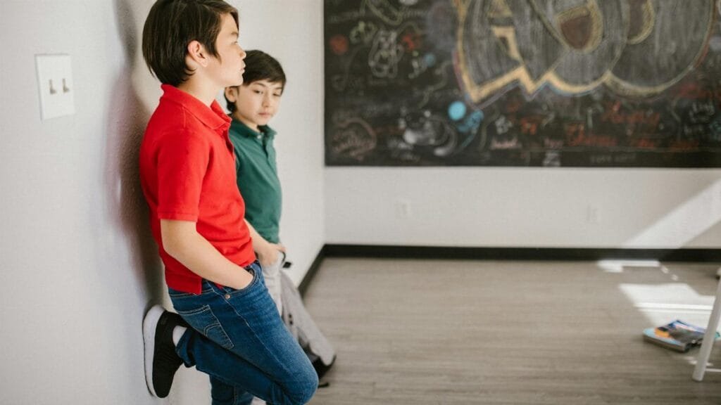 Two boys leaning against a wall in a classroom with graffiti board.