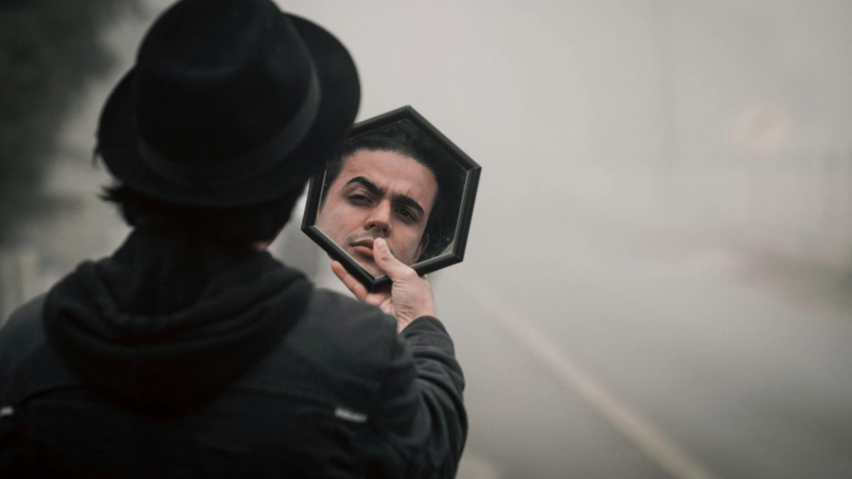 A mysterious man in a black hat reflects in a mirror on a foggy road.
