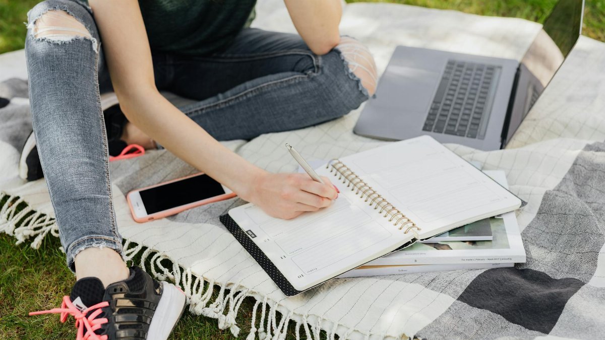 Crop faceless female student in casual clothes taking notes in planner while sitting on blanket with smartphone and laptop on green park grass on sunny day