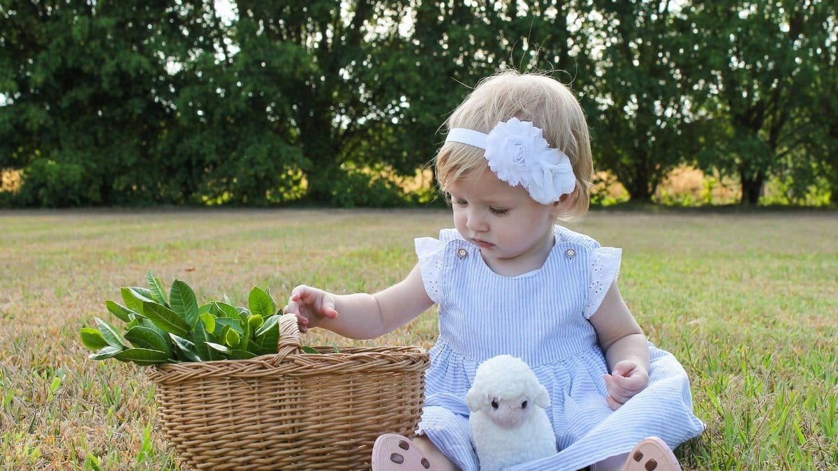 A cute toddler in a striped dress sits outdoors with a basket and plush toy.