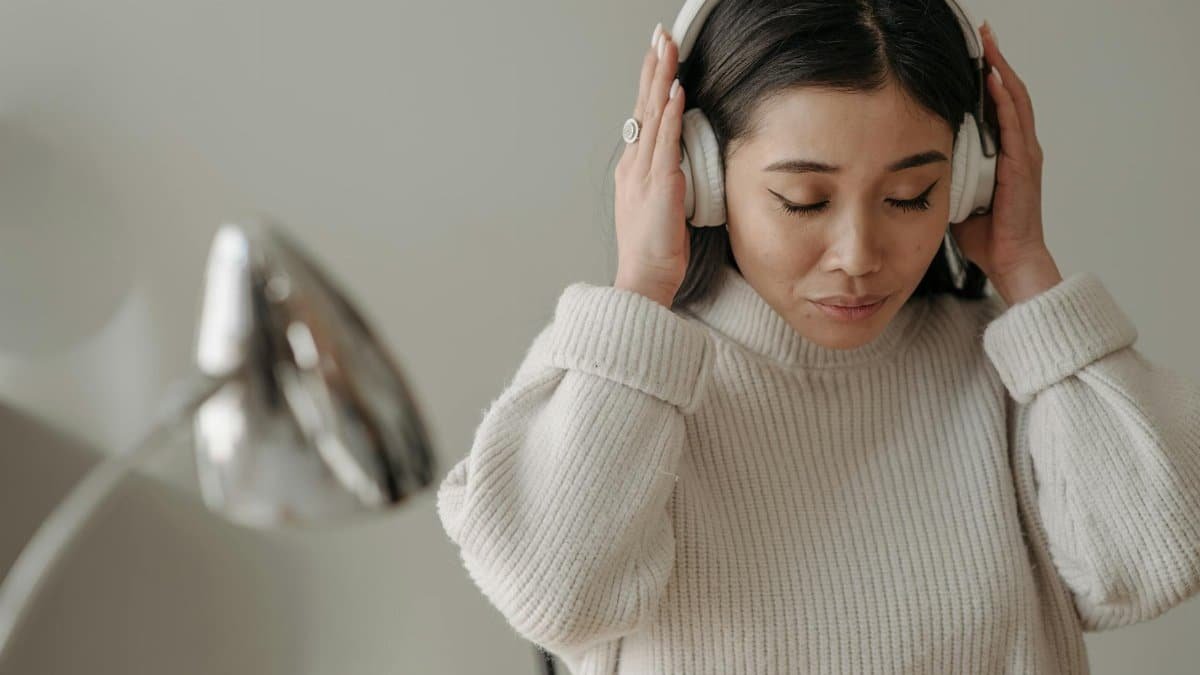 Asian woman wearing headphones and a knitted sweater, enjoying music indoors.
