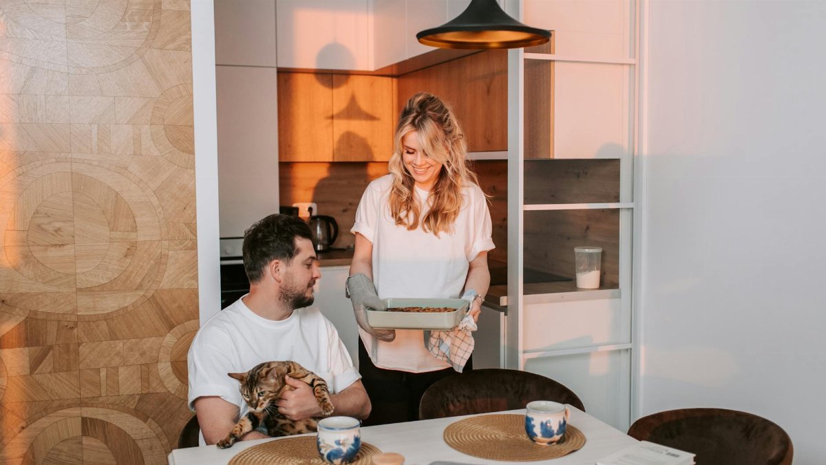 A joyful couple at home enjoying dinner with their pet cat in a cozy kitchen.