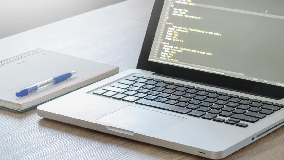 Laptop with code editor open, notebook and pen on wooden desk, perfect for tech and work themes.