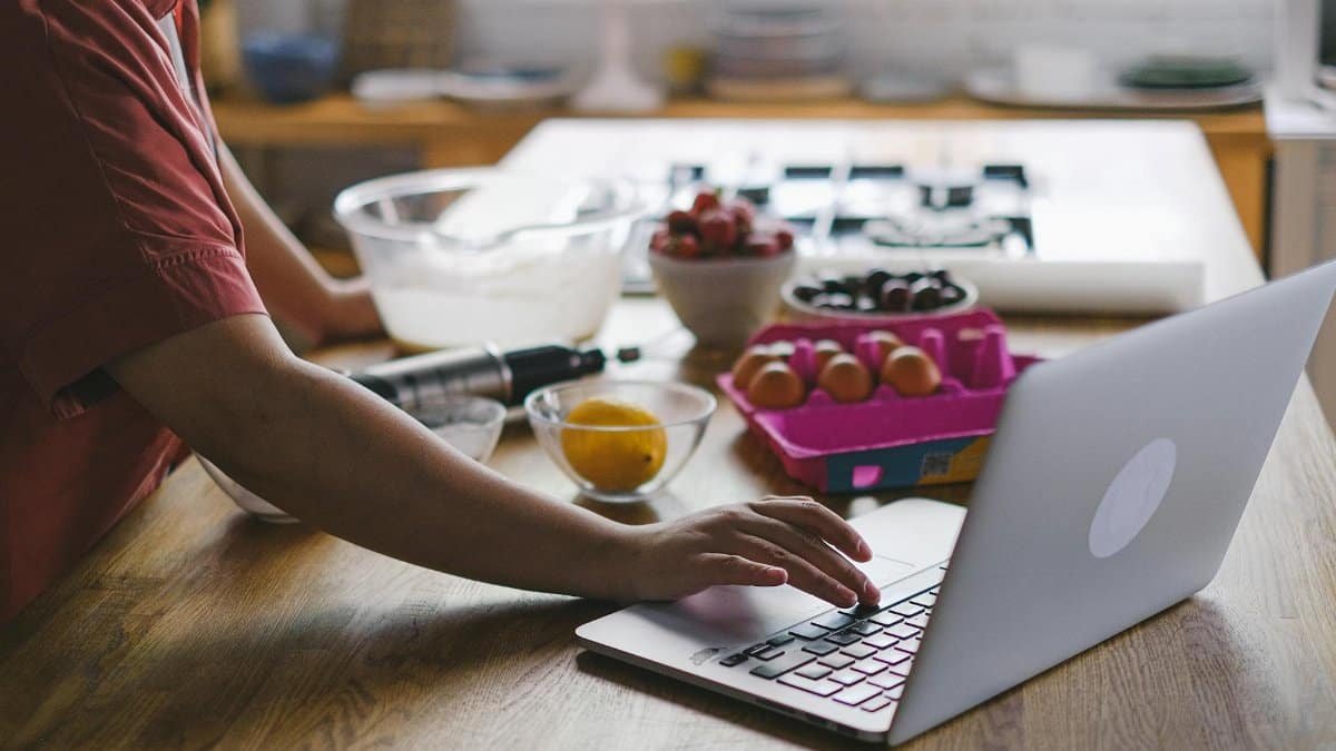 Person using a laptop in the kitchen while preparing baking ingredients. Modern multitasking lifestyle.