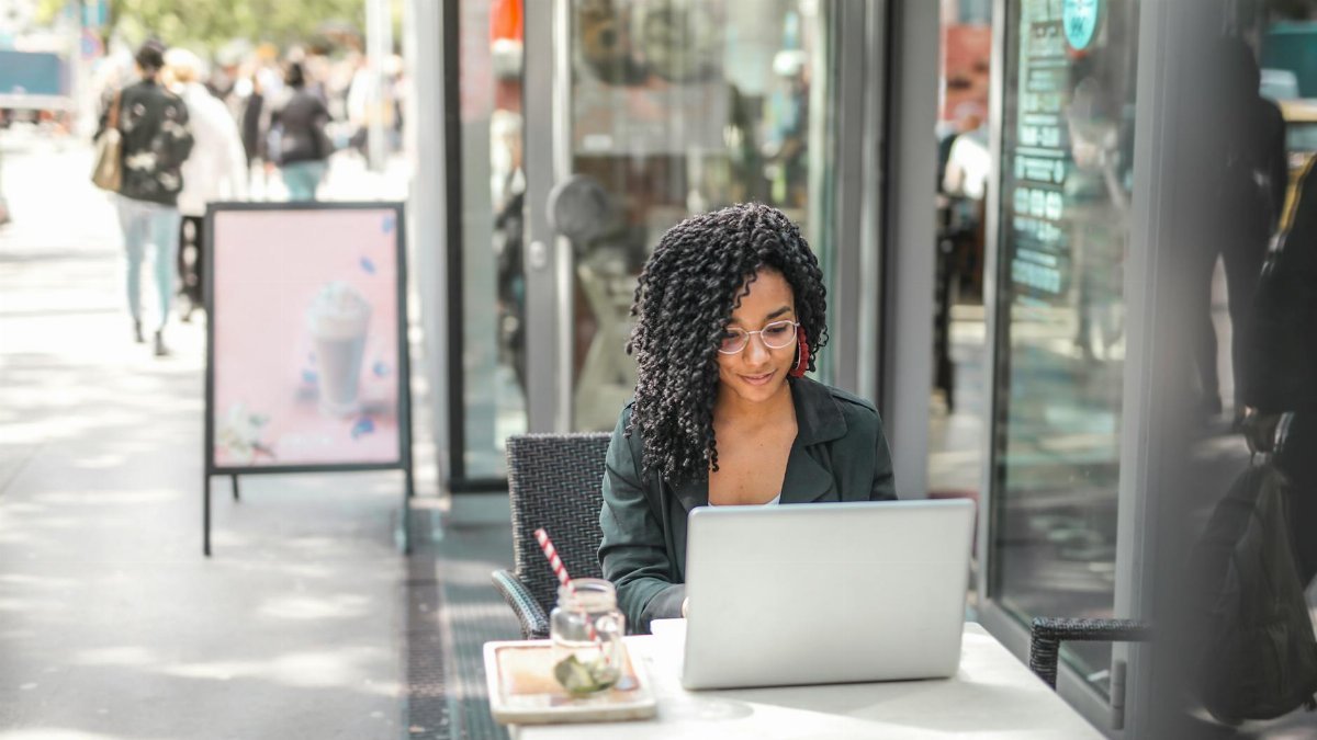 High angle of pensive African American female freelancer in glasses and casual clothes focusing on screen and interacting with netbook while sitting at table with glass of yummy drink on cafe terrace in sunny day