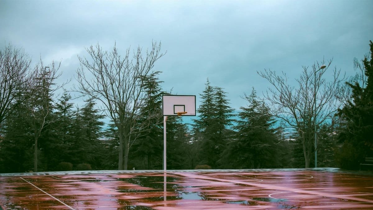 A scenic shot of a drenched basketball court after rain, surrounded by leafless trees.