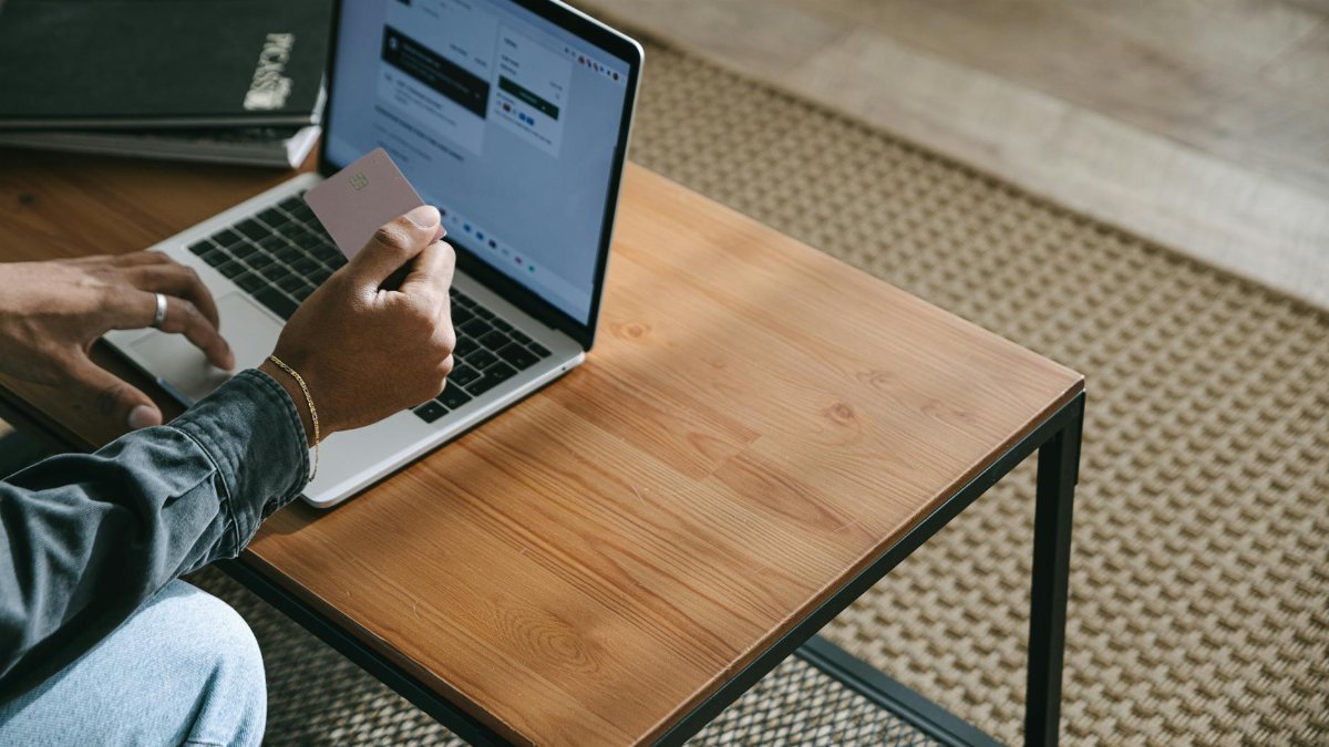 Person holding a credit card while shopping online using a laptop on a wooden table.