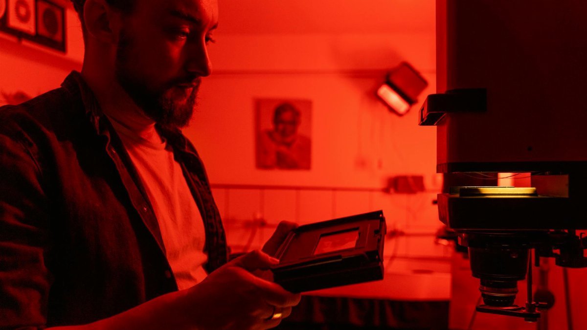 A professional photographer using equipment in a red-lit darkroom, focused on analog photography development.