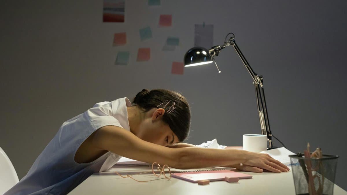 A tired woman rests her head on an office desk under a lamp, symbolizing workplace fatigue.