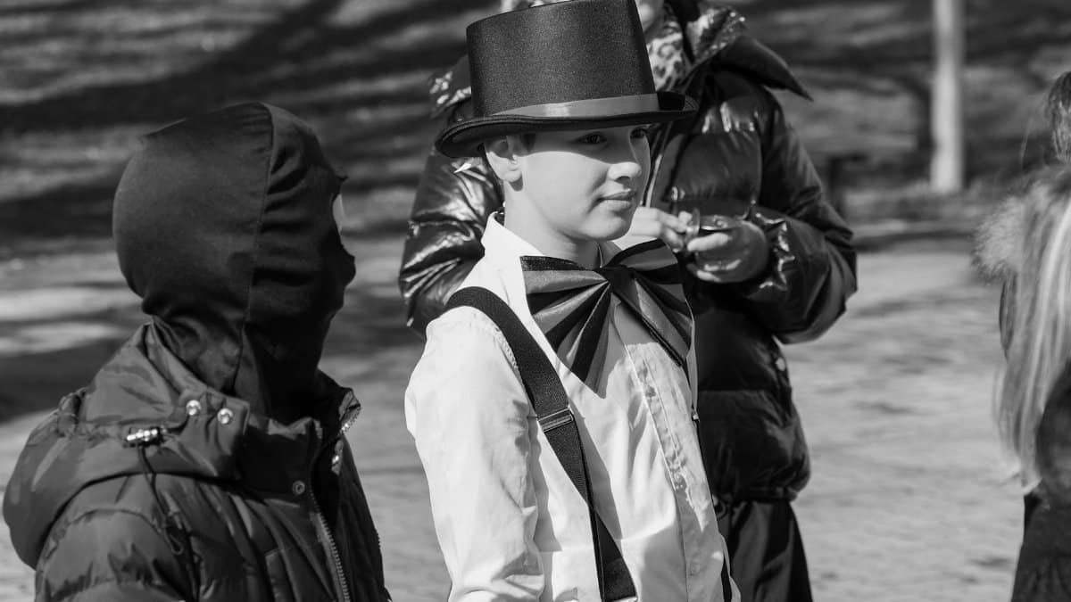 A young performer dressed elegantly with a top hat and bow tie interacts with others on the street.