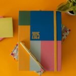 Flat lay of a vibrant 2025 planner with a pen and green plant on yellow background.