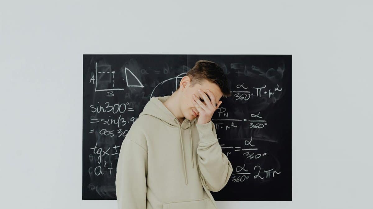 Teen boy covers face in frustration in front of a chalkboard filled with math equations, reflecting academic stress.