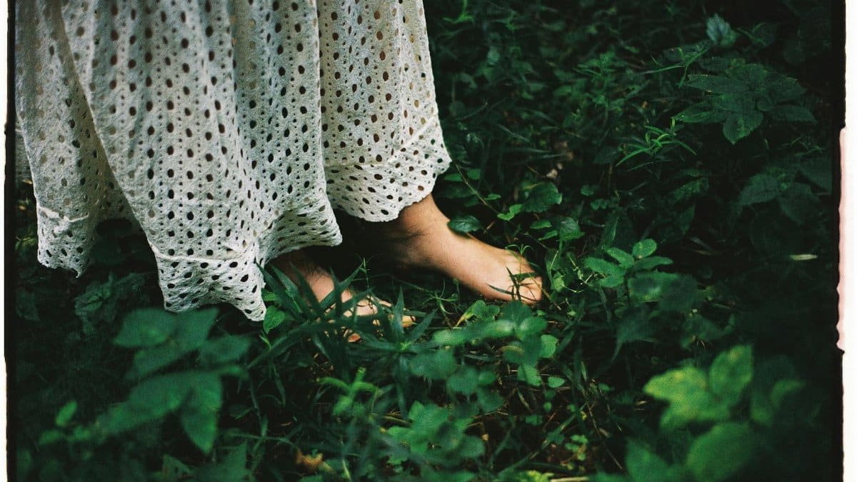 A woman stands barefoot on lush greenery, wearing a white lace dress, portraying harmony and relaxation in nature.