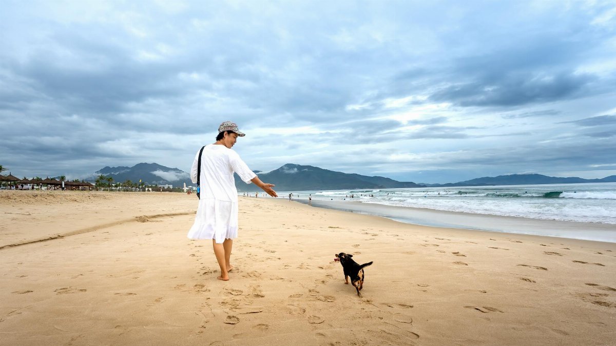 A woman in a white dress walks her dog on a sandy beach under a cloudy sky.