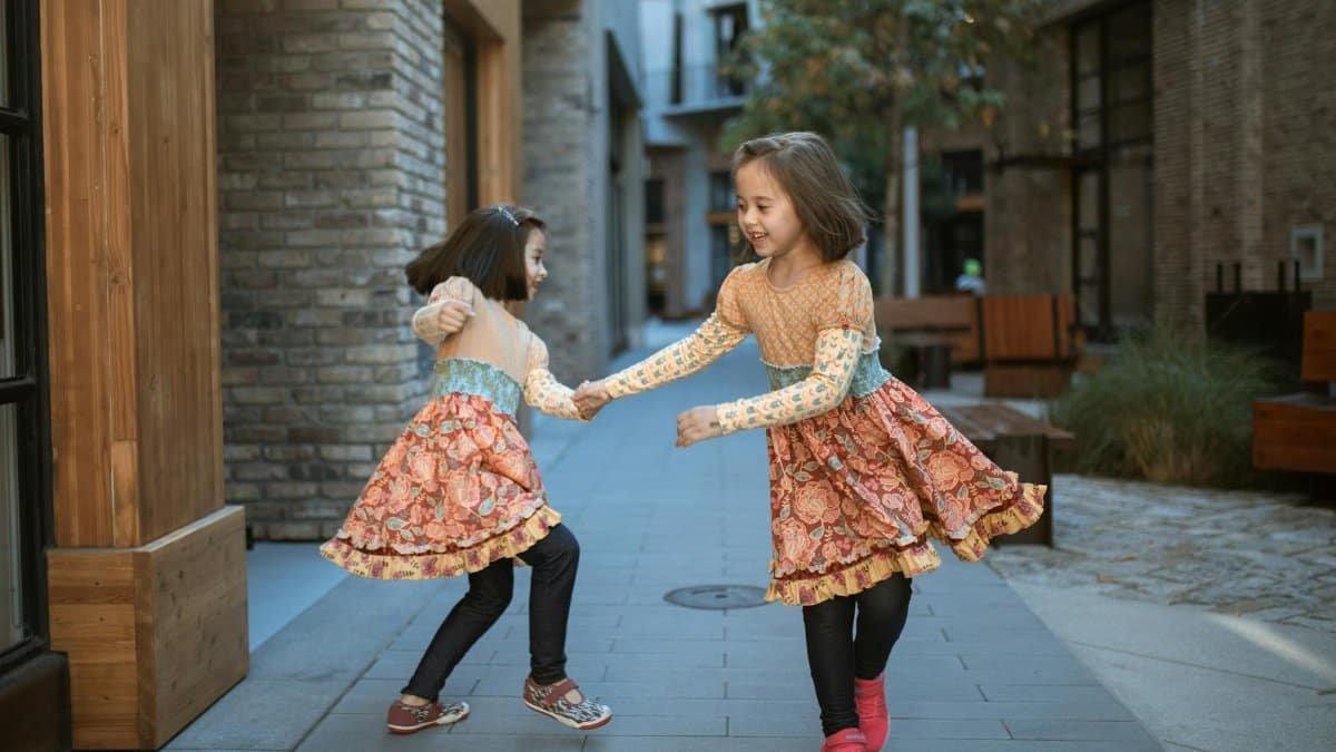 Two Asian girls in colorful dresses enjoy a playful moment on a city sidewalk, embracing childhood joy.