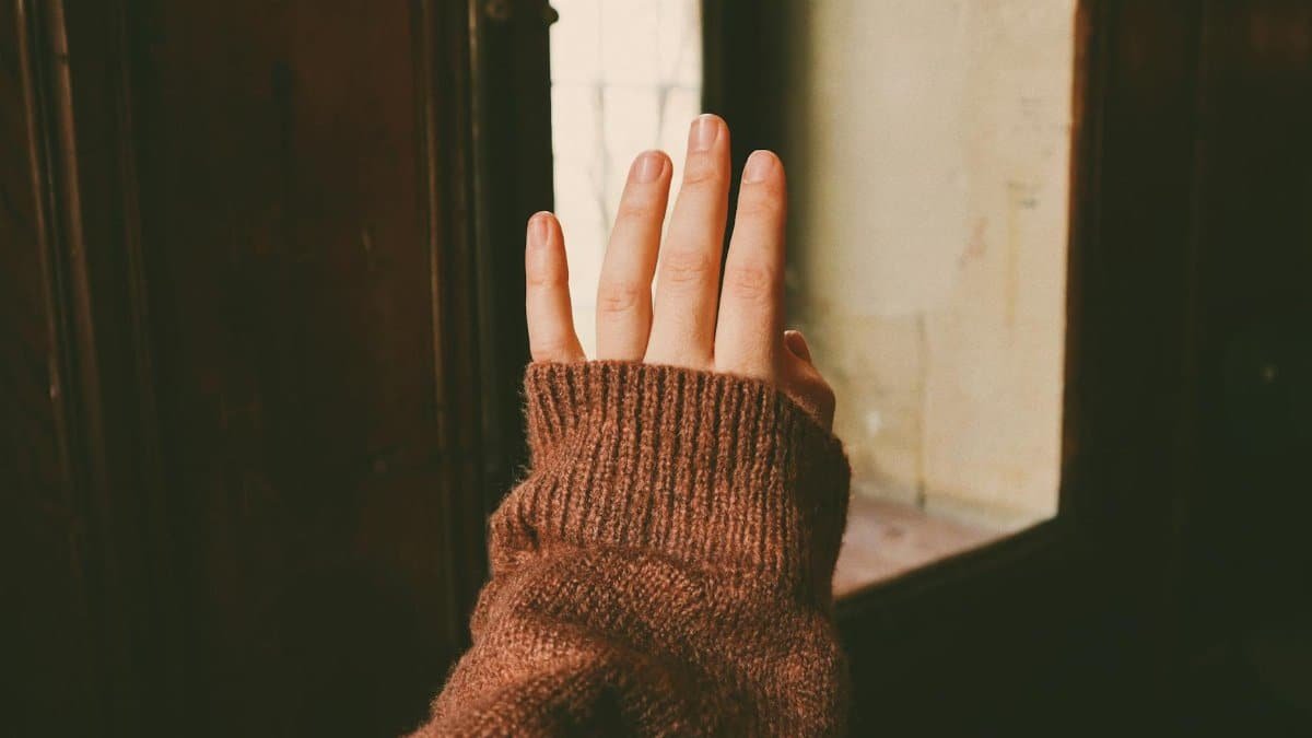 A hand wearing a wool sweater reaching towards a window, featuring warm tones and soft textures.