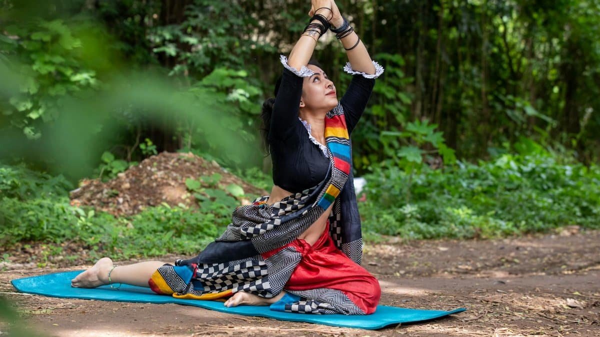 Woman practicing yoga pose outdoors in vibrant traditional attire, surrounded by lush greenery.