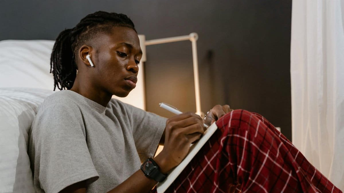 Young man in pajamas writing in a notebook at home, focused and relaxed.