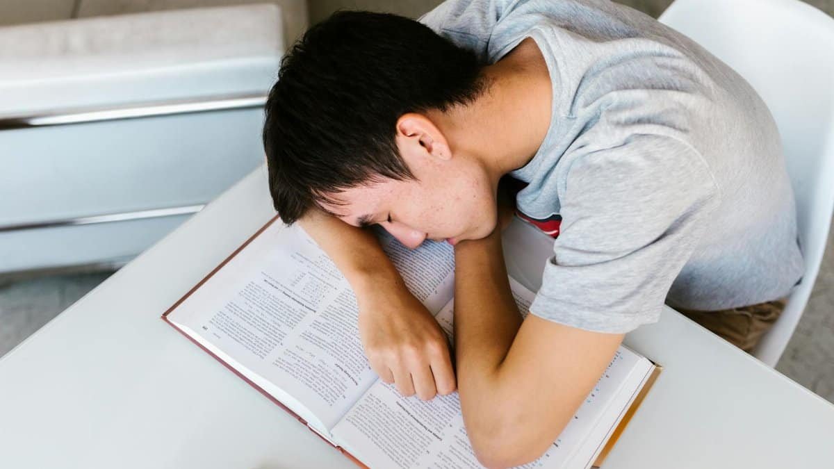 A young man sleeping on a white table with an open textbook, depicting study fatigue.