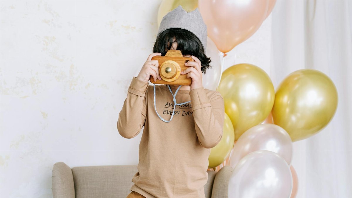 A child wearing a crown uses a toy camera amidst balloons at an indoor birthday party.