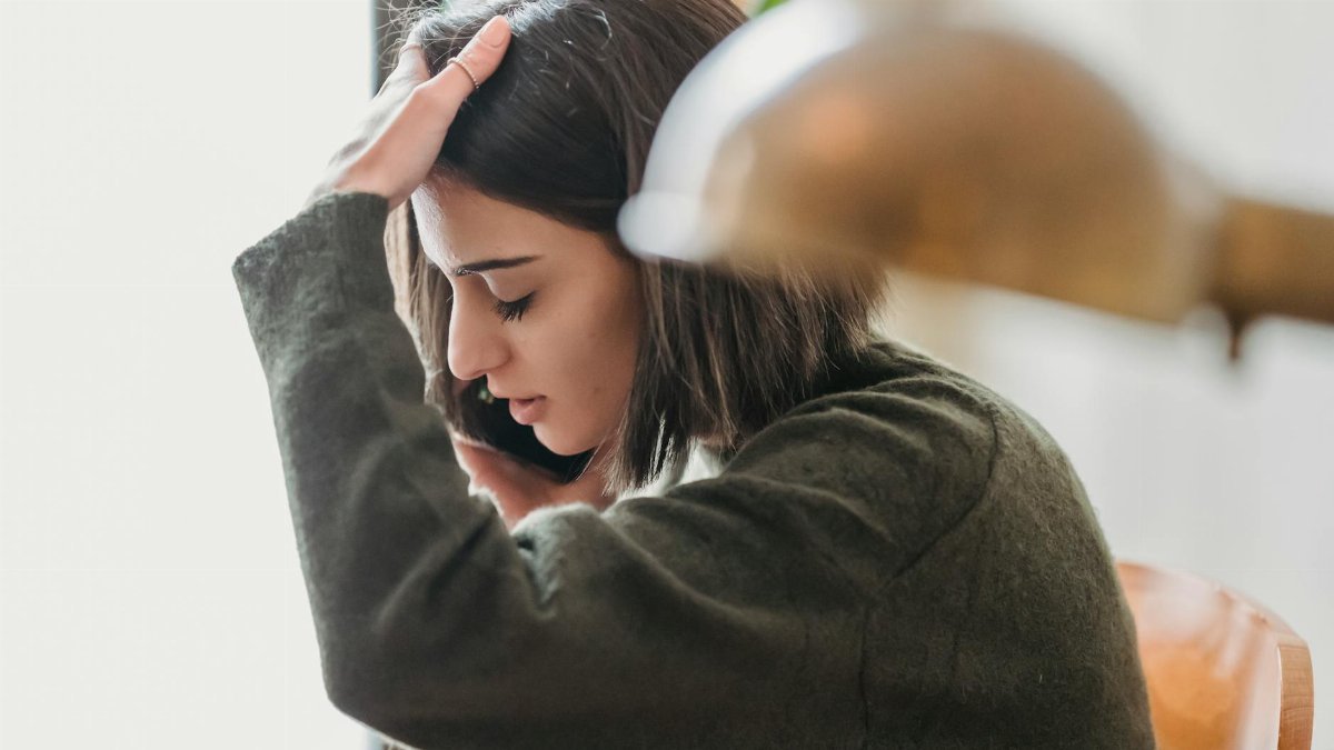 Side view of upset young ethnic female millennial with dark hair grabbing head with closed eyes while having phone conversation sitting on chair at home