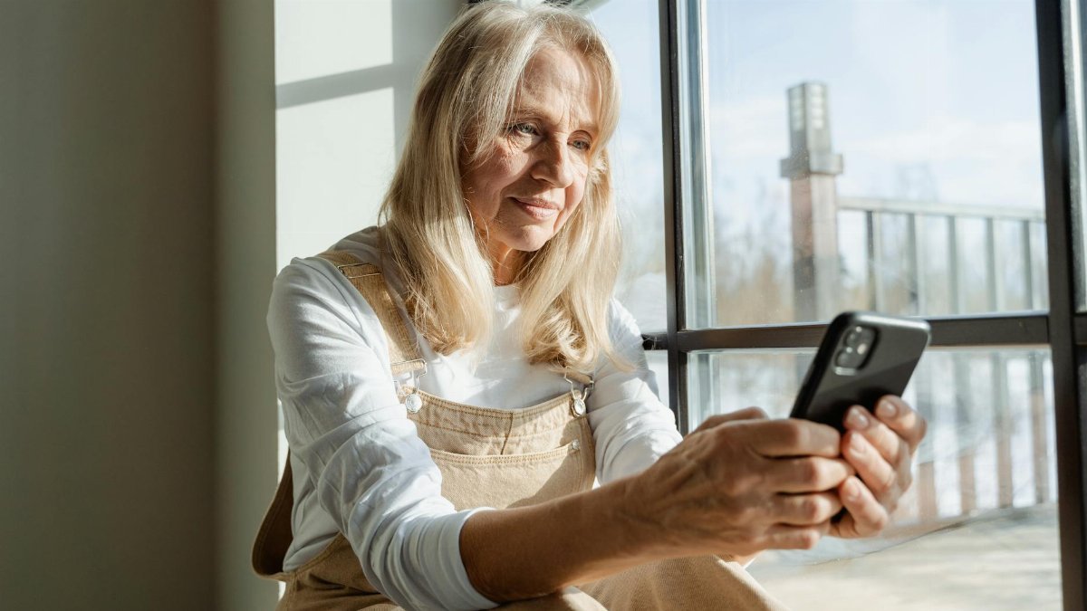 Elderly woman using a smartphone by a window, enjoying leisure time indoors.