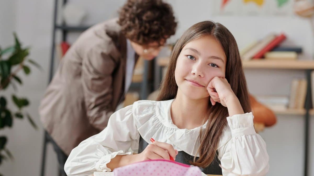 Girl daydreaming at school desk with teacher in background. Education and learning environment.