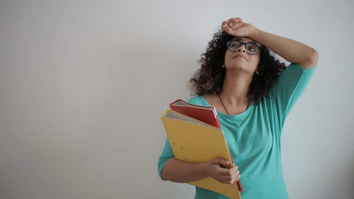 A woman in a teal top looks overwhelmed while holding colorful folders, symbolizing workplace stress.